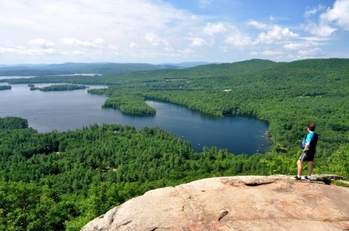 A person stands on a rocky outcrop, overlooking a lush green landscape with lakes and mountains in the distance.