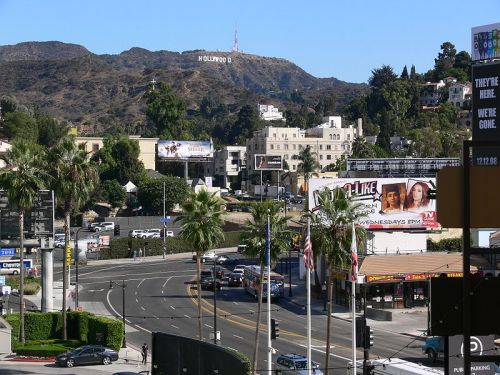 View of Hollywood sign on a hillside, with palm trees and busy streets below, featuring various billboards and buildings.