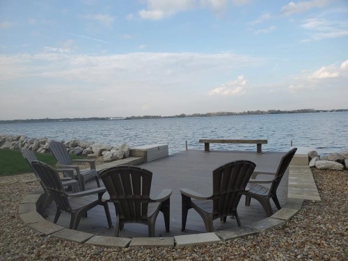 A serene lakeside view featuring a wooden deck with Adirondack chairs and a stone border, under a cloudy sky.