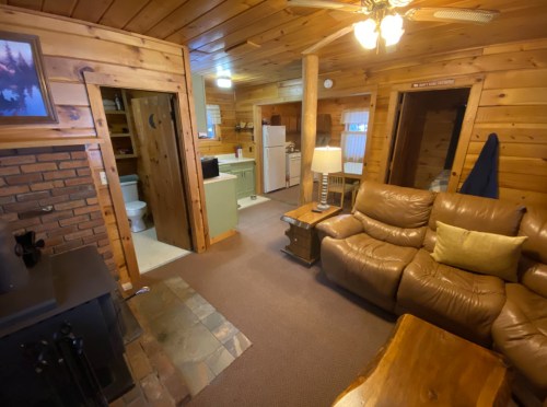 Cozy wooden cabin interior featuring a living area, kitchen, and bathroom, with leather furniture and rustic decor.