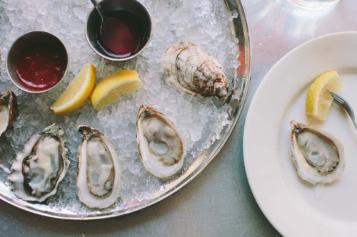 A platter of oysters on ice with lemon wedges and dipping sauces, alongside a plate with a single oyster and lemon.