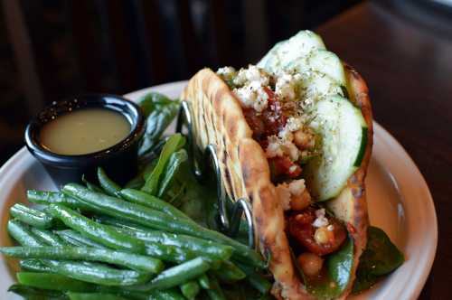 A pita filled with vegetables and feta cheese, served with green beans and a small bowl of dressing on the side.