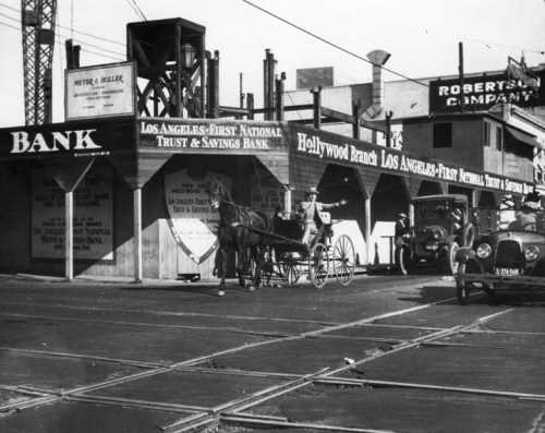 Historic street scene featuring a horse-drawn carriage, early cars, and bank buildings in Los Angeles.