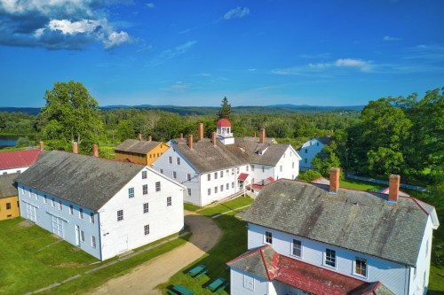 Aerial view of historic white buildings with red roofs, surrounded by green trees and mountains under a blue sky.