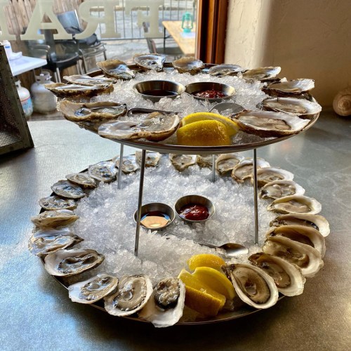A two-tiered platter of fresh oysters on ice, garnished with lemon wedges and dipping sauces.