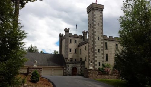 A large stone castle with a tower, surrounded by trees and a driveway leading to the entrance.