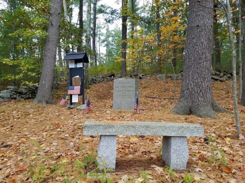 A stone bench in a wooded area near a memorial with flags, surrounded by autumn leaves and trees.