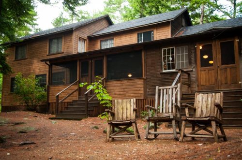 A rustic wooden cabin surrounded by trees, featuring a porch and rocking chairs in front.
