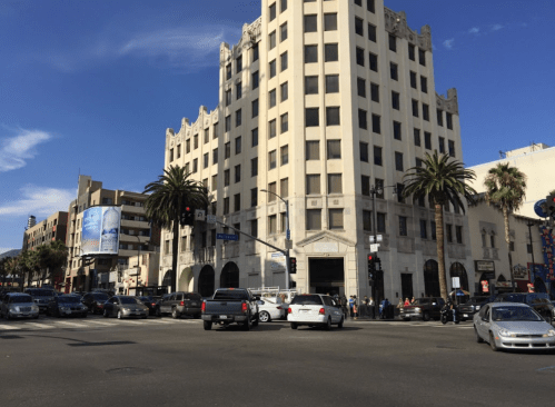 A busy intersection featuring a tall, historic building, palm trees, and various vehicles on a sunny day.