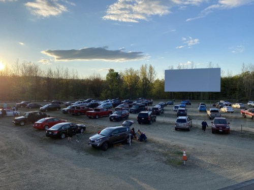A drive-in movie theater with parked cars and a large blank screen under a clear sky at sunset.