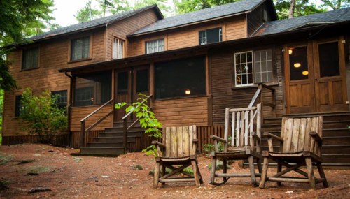 A rustic wooden cabin surrounded by trees, featuring a porch and rocking chairs in front on a gravel path.