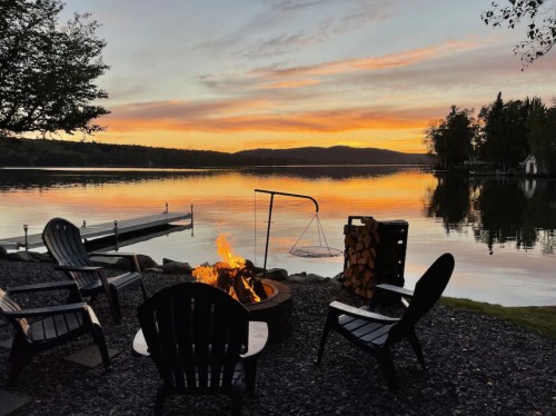 A serene lakeside scene at sunset, featuring a fire pit, chairs, and a calm water reflection.