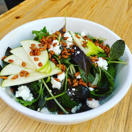 A fresh salad with mixed greens, sliced apples, goat cheese, and crunchy seeds in a white bowl on a wooden table.