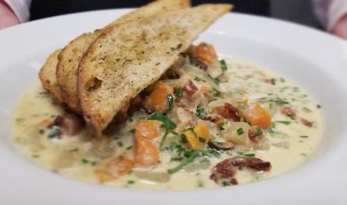 A creamy soup with vegetables and bacon, garnished with herbs, served with toasted bread on top.