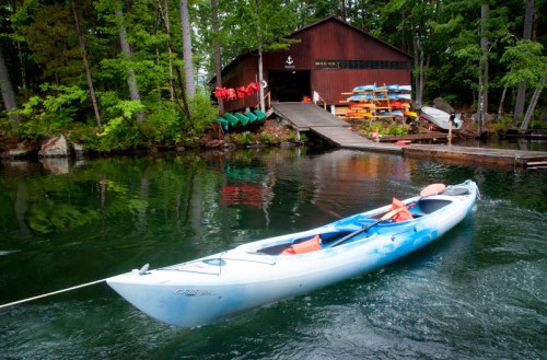 A kayak floats in calm water near a boathouse surrounded by trees, with colorful kayaks and canoes on the dock.