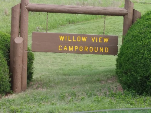 Wooden sign reading "Willow View Campground" hanging between two green bushes in a grassy area.