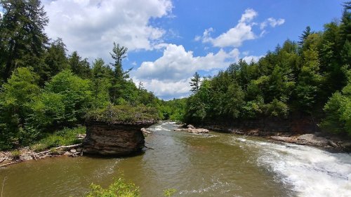 A serene river flows through a lush green landscape, with rocky formations and a blue sky dotted with clouds.