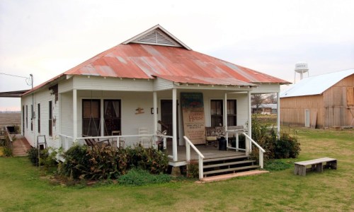 A quaint white house with a rusty red roof, surrounded by greenery and a wooden bench in front.