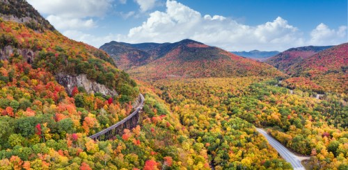 Aerial view of vibrant autumn foliage covering rolling hills and a winding road under a blue sky with fluffy clouds.
