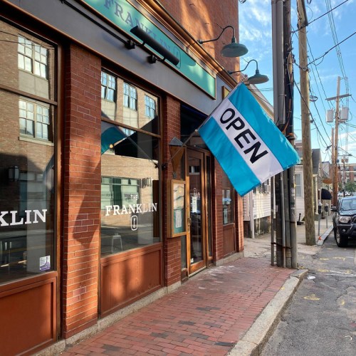 A storefront with a blue and white "OPEN" flag, brick sidewalk, and large windows reflecting the sky.