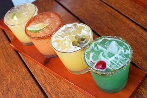 Four colorful cocktails on a wooden tray, featuring lime and cherry garnishes, served over ice.