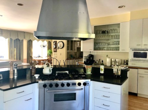 Modern kitchen with a large island, stainless steel hood, gas stove, and glass-front cabinets filled with dishes.