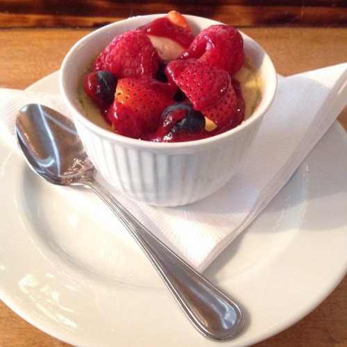 A small white bowl filled with creamy dessert topped with fresh berries and a drizzle of sauce, placed on a plate with a spoon.