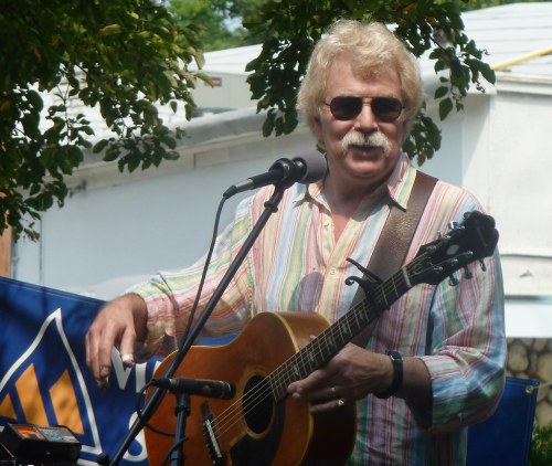 A musician with a mustache plays an acoustic guitar and sings at an outdoor event, wearing a colorful striped shirt.