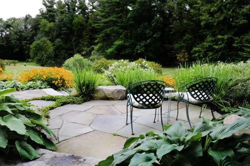 A serene garden scene featuring two green chairs on a stone patio surrounded by vibrant flowers and lush greenery.