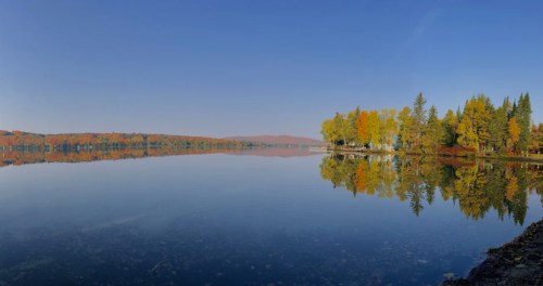 A serene lake reflecting colorful autumn trees under a clear blue sky.