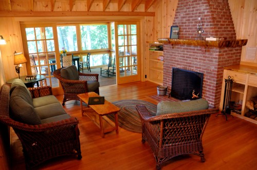 Cozy living room with wicker furniture, a brick fireplace, and large windows overlooking a wooded area.