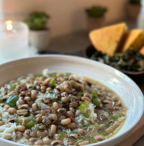 A bowl of rice topped with black-eyed peas and green onions, served with cornbread and greens in the background.