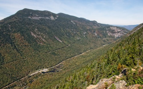 Aerial view of a lush green valley surrounded by mountains, with a winding road and a small body of water below.