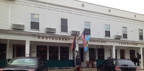 Exterior of a restaurant with signs for "Chocolate" and "Confections," featuring flags and parked cars in front.