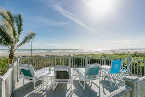 A sunny beach view from a deck with lounge chairs overlooking the ocean and palm trees swaying in the breeze.