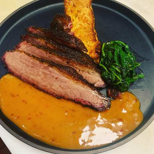 A plate featuring sliced brisket, a piece of toasted bread, sautéed greens, and a rich sauce.