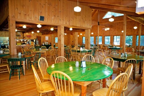 A spacious wooden dining area with green and natural wood tables and chairs, featuring large windows and warm lighting.