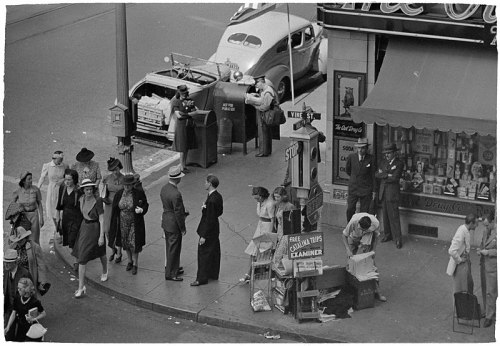 A busy street scene from the past, featuring pedestrians, a vintage car, and a newsstand with people interacting.