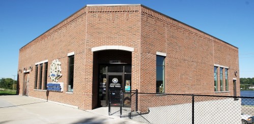 A brick building with large windows and a sign, located near a body of water, surrounded by a fence.