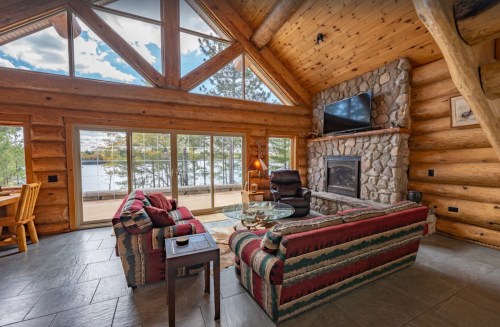 Cozy log cabin interior with large windows, stone fireplace, and comfortable seating overlooking a lake.