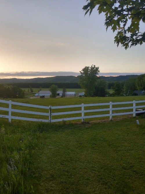 A serene landscape featuring a green field, white fence, and distant mountains under a sunset sky.
