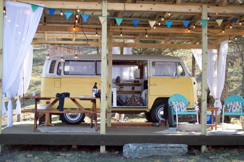 A vintage yellow van parked under a wooden shelter, surrounded by string lights and colorful bunting.