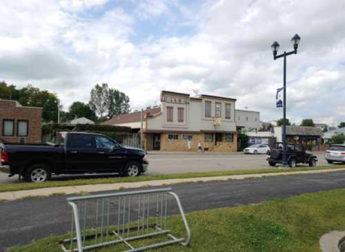 A street view of a small building with a sign, surrounded by parked cars and a grassy area. Cloudy sky above.