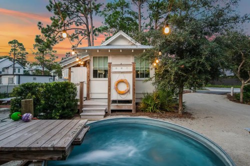 Charming cottage with a hot tub, surrounded by trees and outdoor lighting, at sunset.