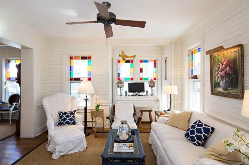 Cozy living room with white furniture, colorful stained glass windows, and a ceiling fan, featuring a small desk and decor.
