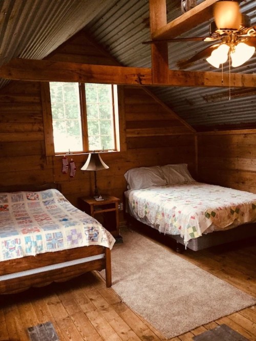 Cozy cabin interior with two beds, wooden walls, a lamp, and a large window letting in natural light.