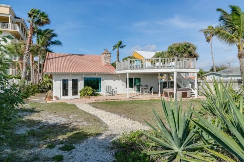 A beach house with a deck, surrounded by palm trees and a grassy area, under a clear blue sky.