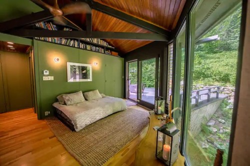 Cozy bedroom with green walls, wooden ceiling, large windows, and a view of nature, featuring a bed and decorative lanterns.