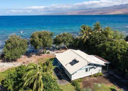 Aerial view of a coastal house surrounded by palm trees and clear blue water, with mountains in the background.