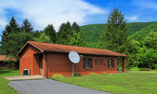 A brick building with a satellite dish, surrounded by green trees and mountains under a blue sky.
